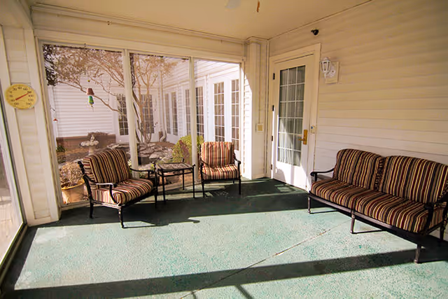 A screened-in porch area with three cushioned chairs and a small glass-top table. The chairs have striped upholstery in shades of brown, beige, and black. The porch has a green carpeted floor and white walls with a door leading outside. Outside the screened area, there are trees and shrubs visible.
