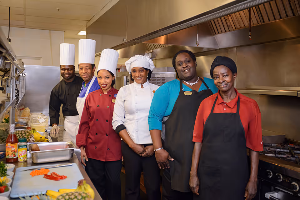 Six kitchen staff in chef uniforms and aprons stand together in a commercial kitchen with prep counters and cooking equipment.