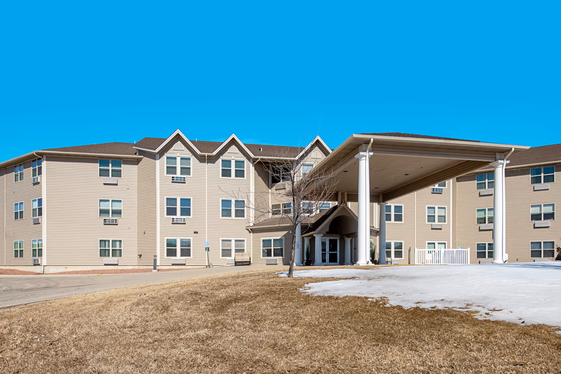 Exterior view of a three-story beige senior living facility building with multiple windows and a large covered entrance supported by white columns. The foreground shows a patchy lawn with some snow and a clear blue sky above.