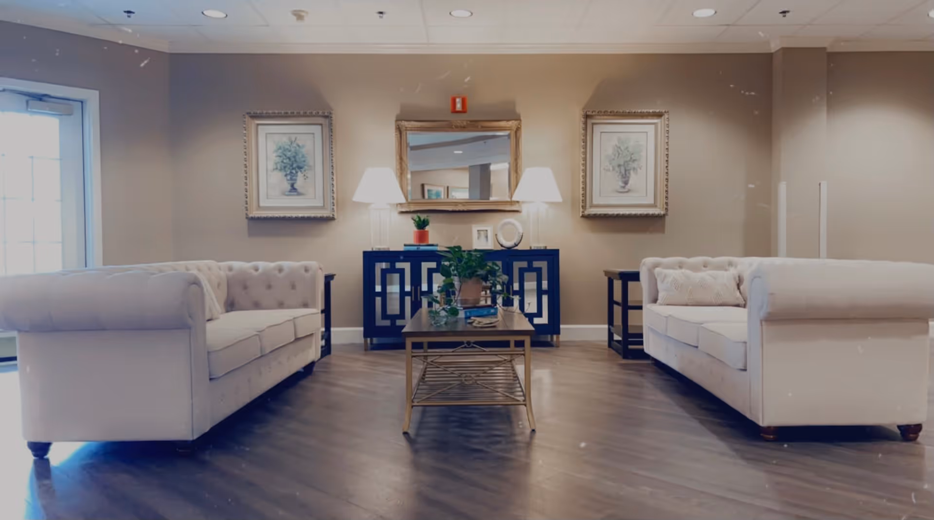 A cozy living room area with two white tufted sofas facing each other, a wooden coffee table with plants and books in the center, a blue console table against the wall with two lamps, decorative items, and a large mirror above it. Two framed botanical prints hang on the wall on either side of the mirror. The room has wooden flooring and neutral-colored walls.