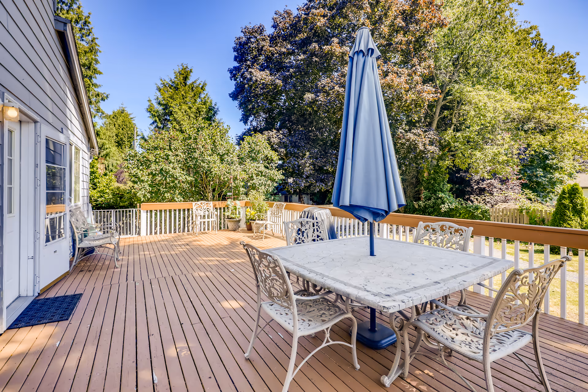 A spacious outdoor wooden deck attached to a building with a white door and windows. The deck features a square marble table with a closed blue umbrella in the center and four ornate metal chairs around it. Additional metal chairs and potted plants are placed along the railing. The background shows lush green trees under a clear blue sky.