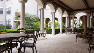 Covered outdoor patio area with multiple round metal tables and chairs, supported by a series of arches and columns. The patio overlooks a landscaped garden with bushes and greenery, adjacent to a multi-story building.
