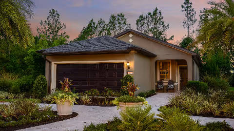 Front exterior of a single-story house with a dark two-car garage, paved walkway and lush landscaping at dusk.
