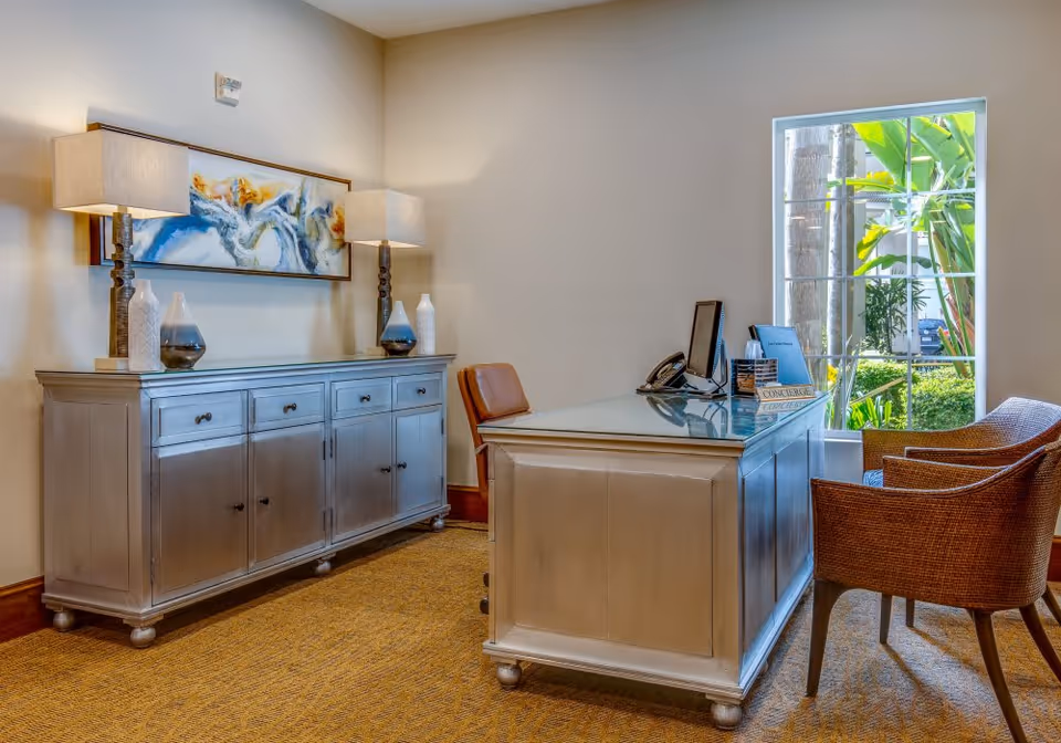 A bright reception-style office with a wooden desk, guest chairs, a sideboard with lamps and decorative vases, and a window showing greenery outside.