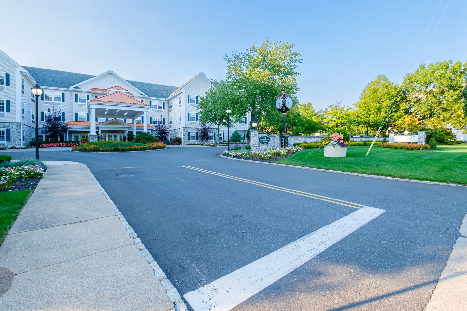 Front exterior view of a senior living facility named Brandywine Summit by Monarch, featuring a large three-story building with white siding and stone accents, a covered entrance with columns, landscaped flower beds, a clock on a stone pedestal, and a paved driveway with a sidewalk.