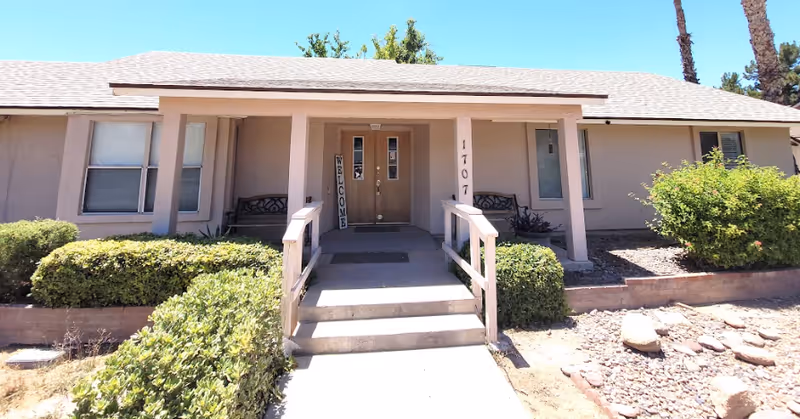 Front entrance of a single-story senior living facility with steps, pillars, and shrubs.