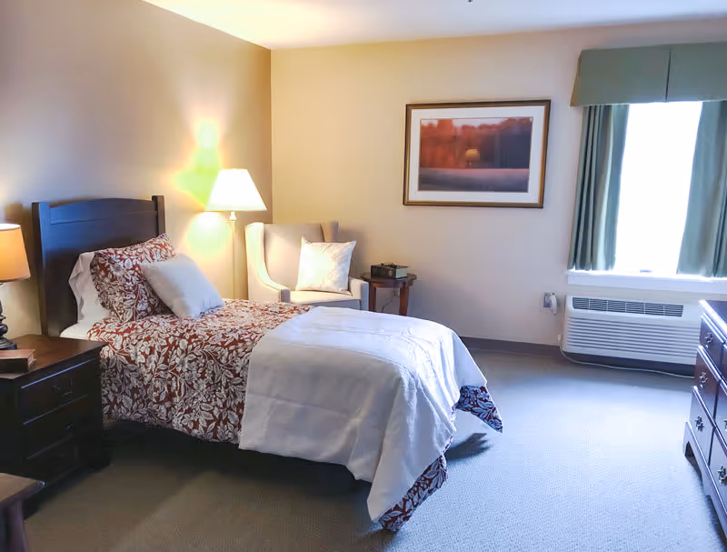 A cozy bedroom in a senior living facility featuring a single bed with floral bedding and white blanket, a cushioned armchair with a pillow, two bedside tables with lamps, a framed landscape picture on the wall, a window with green curtains, and a heating/cooling unit below the window.