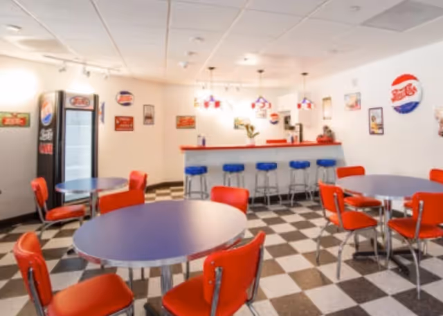 A retro-style dining area with round tables and red chairs, a checkered black and white floor, a counter with blue stools, and vintage soda-themed wall decorations including a Pepsi refrigerator and signs.