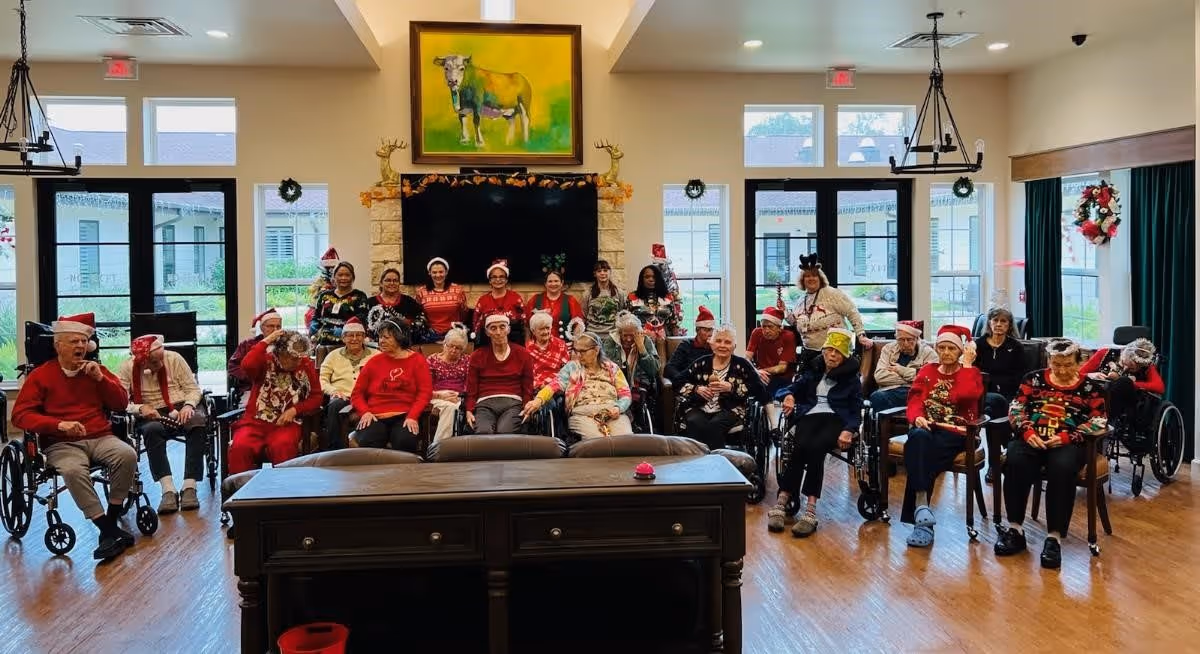A group of elderly residents and staff at The Bradford Memory Care and Assisted Living gathered in a spacious, well-lit common room decorated for the holidays. Many are wearing festive clothing and Santa hats, seated in chairs and wheelchairs facing the camera. The room features large windows, a mounted flat-screen TV above a stone fireplace, and a colorful painting of a cow on the wall.