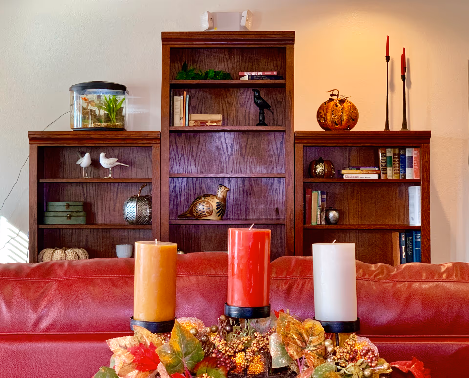 A cozy living room area with a red leather couch in the foreground. Behind the couch are three wooden bookshelves filled with decorative items such as books, small sculptures, candles, and a small fish tank. The room is warmly lit with a neutral wall background.