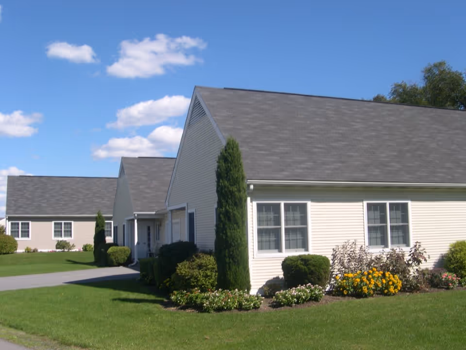 Exterior view of single-story residential buildings with beige siding and gray roofs under a blue sky with scattered clouds. The buildings are surrounded by well-maintained green lawns, shrubs, and flower beds with yellow and pink flowers.