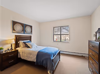 A neatly arranged bedroom with a single bed featuring white bedding and a blue throw blanket. There is a wooden nightstand with a lamp and decorative items on the left side of the bed. Above the bed, two framed circular art pieces are hung on the beige wall. A window with a view of trees and buildings is centered on the far wall. On the right side of the room, there is a wooden dresser with a mirror and some decorative items on top. The room has beige carpet and light-colored walls.