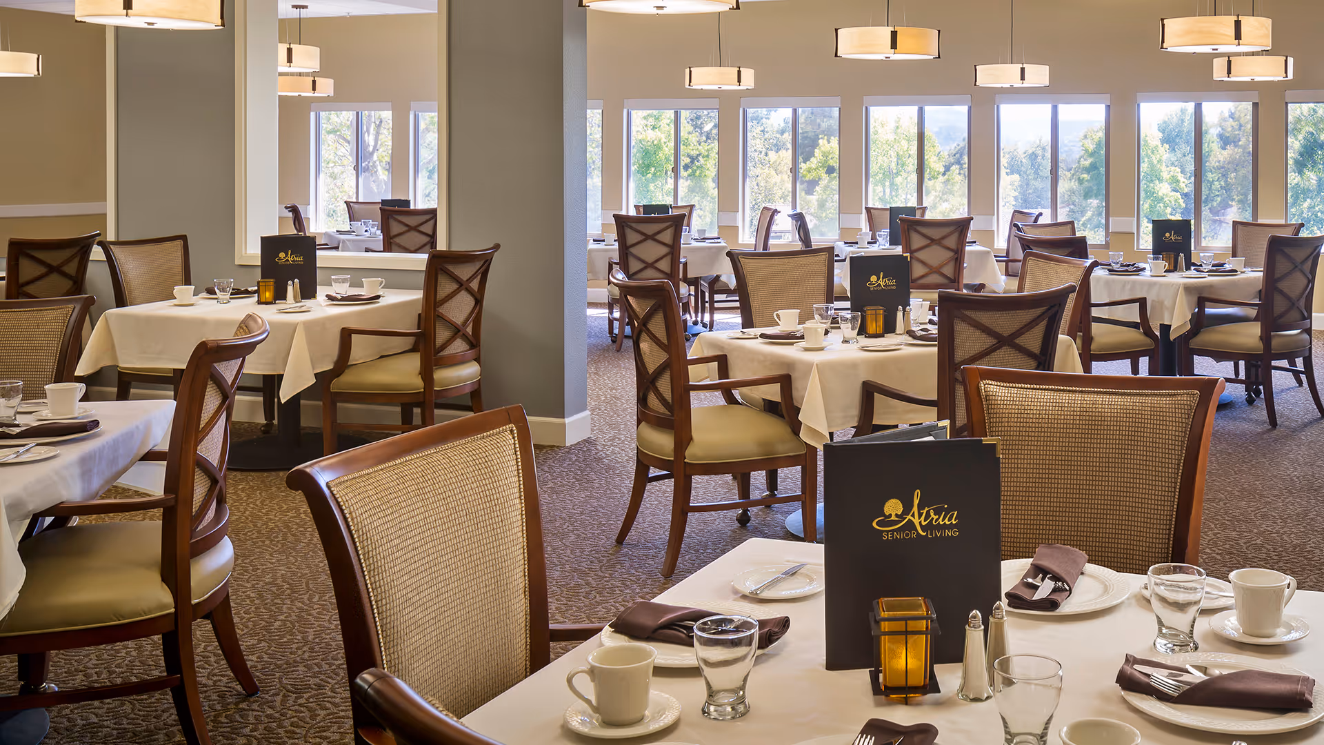 A bright and spacious dining room with multiple tables covered in white tablecloths, each set with cups, glasses, plates, and silverware. The chairs have wooden frames with cushioned seats and backs. Menus labeled 'Atria Senior Living' are placed on several tables. Large windows in the background let in natural light and offer views of greenery outside.