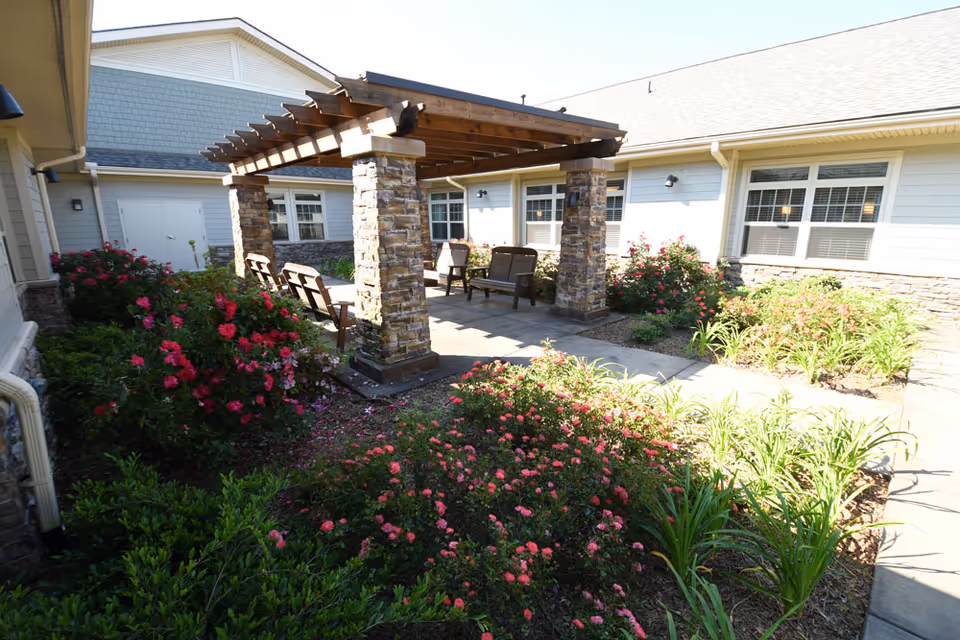 Outdoor courtyard area at Oakview Park featuring a wooden pergola with stone pillars, several chairs and benches underneath, surrounded by blooming pink and red flowers and green plants, with the building's exterior walls and windows visible in the background.