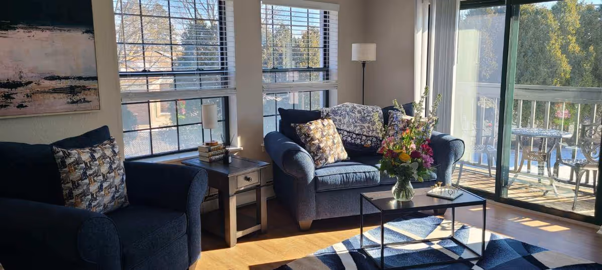 Sunlit living room with blue upholstered sofa and armchair, a coffee table with a vase of flowers, large windows and a sliding glass door opening to a balcony.
