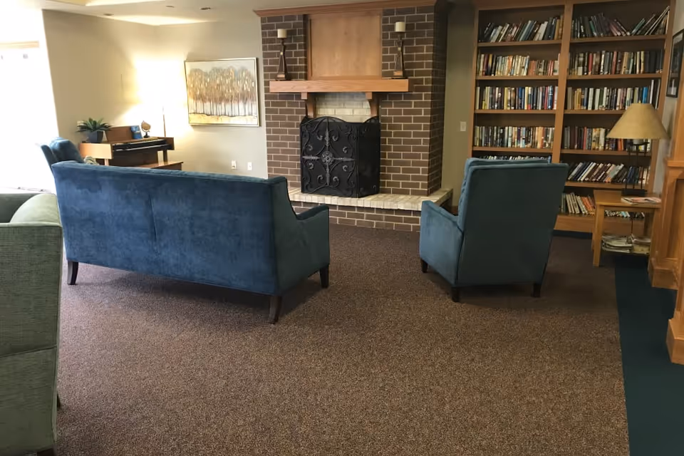 A cozy living room area with a blue sofa and matching armchair facing a brick fireplace with a decorative black metal screen. To the right, there is a wooden bookshelf filled with books and a floor lamp. On the left side, a piano and a framed artwork are visible, with a window letting in natural light.