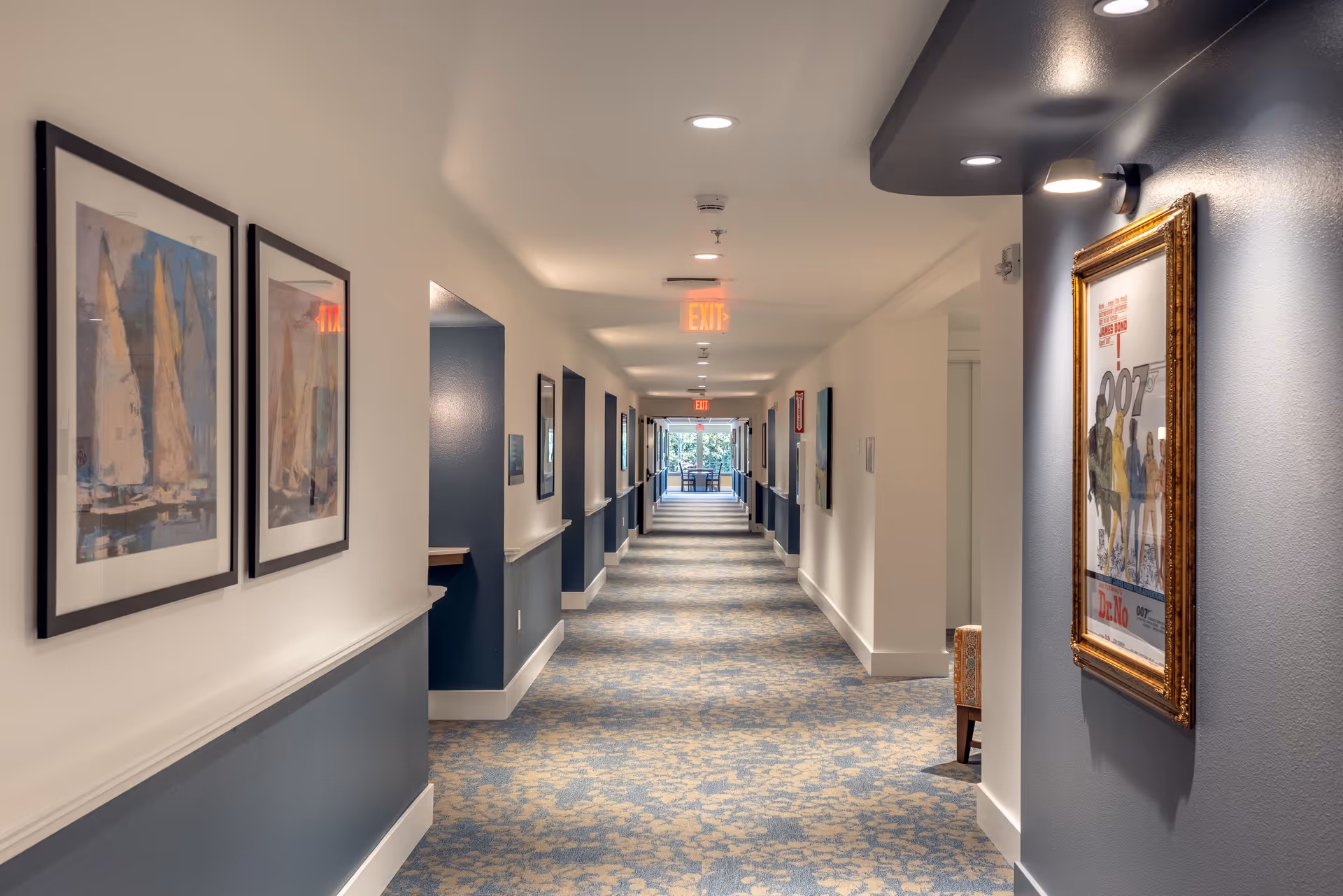 A long, well-lit hallway in a senior living facility with blue and beige patterned carpet, white walls with blue lower panels, framed artwork on the walls, and exit signs on the ceiling. At the far end, there is a seating area near a window with natural light.