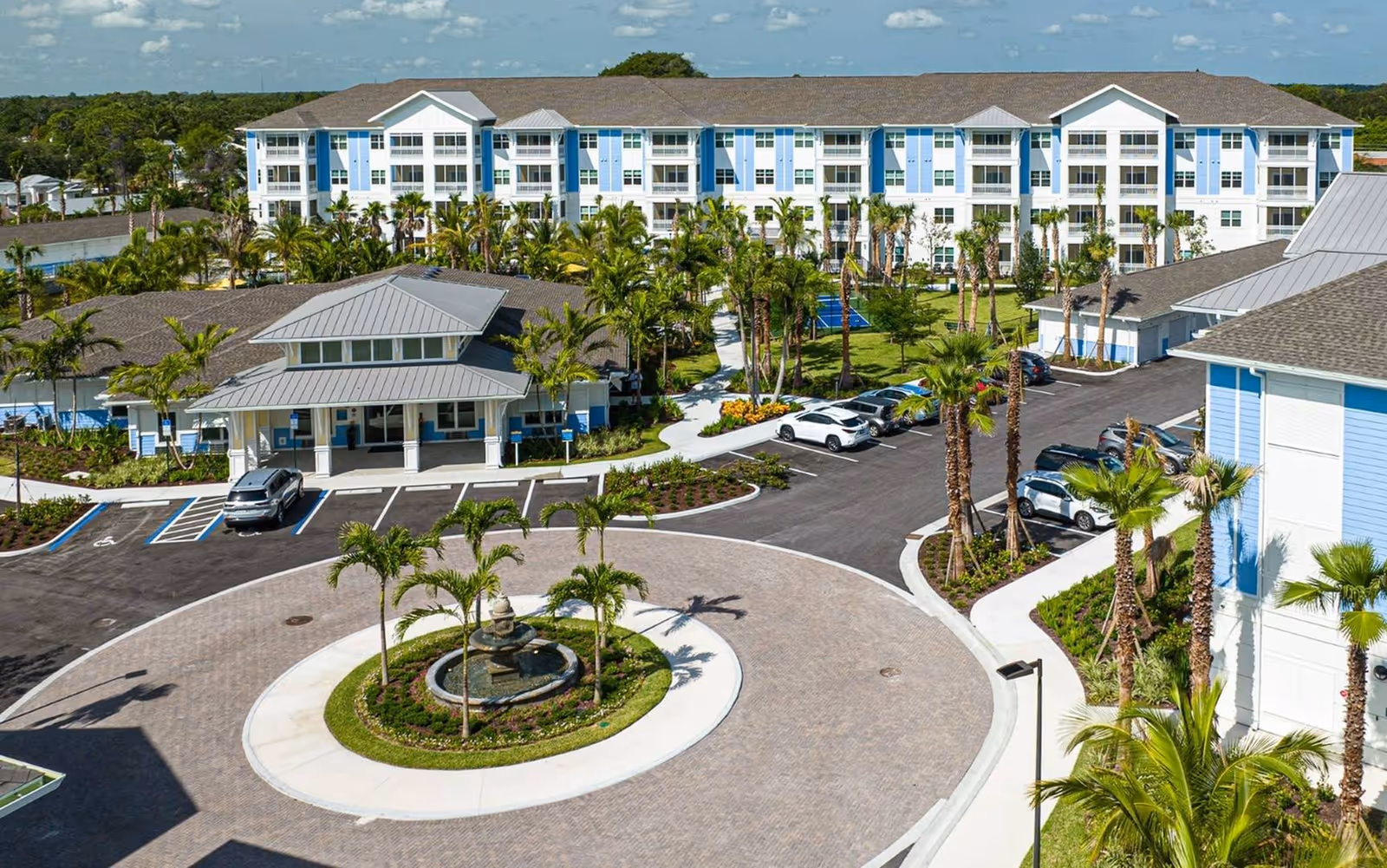 Aerial view of The Sovana At Stuart's landscaped front with a circular fountain, palm trees, parking area, and multi-story light-blue residential buildings.