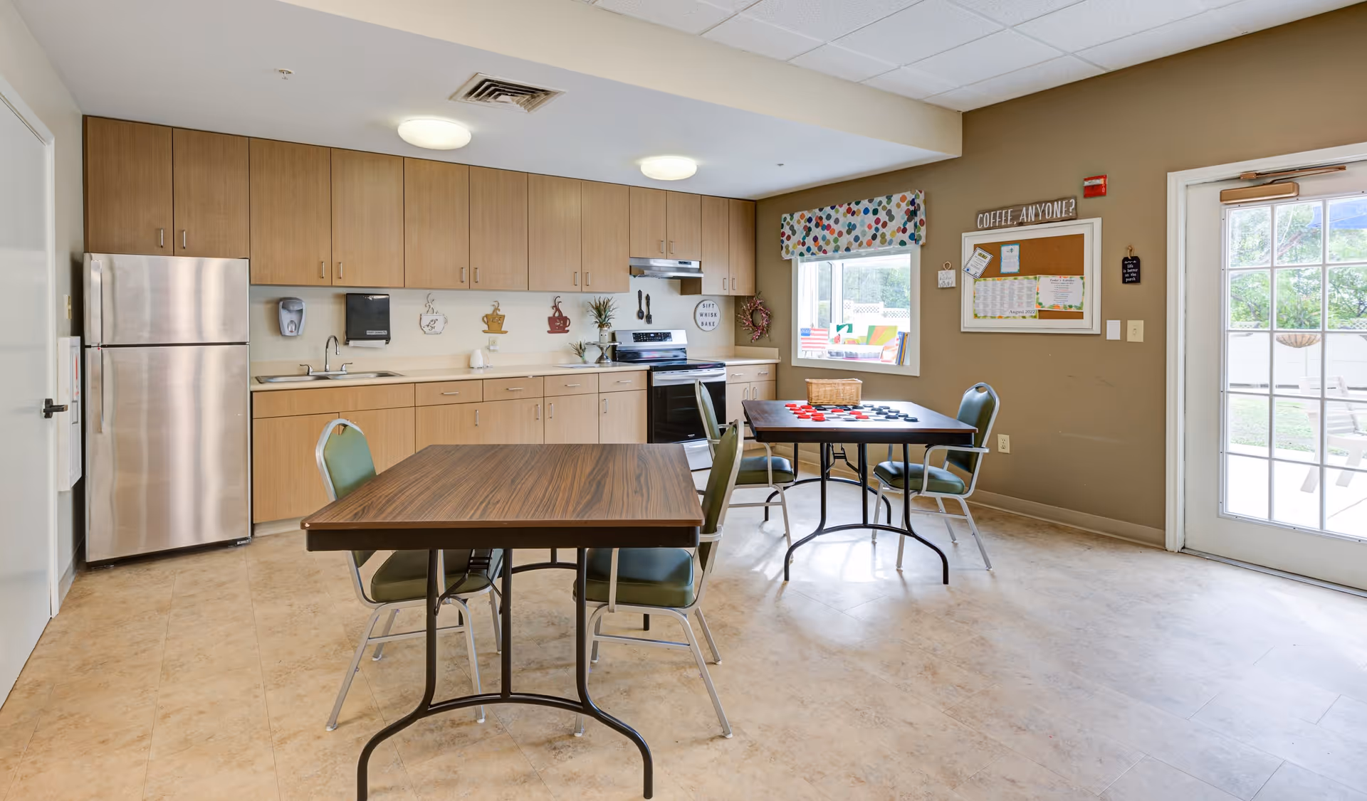 A bright kitchen and dining area in a senior living facility with wooden cabinets, a stainless steel refrigerator, stove, and sink. Two tables with green chairs are set up, one with a checkers game on it. A window with a colorful valance and a bulletin board with the sign 'COFFEE, ANYONE?' are visible on the beige walls. A glass door leads outside to a patio area.