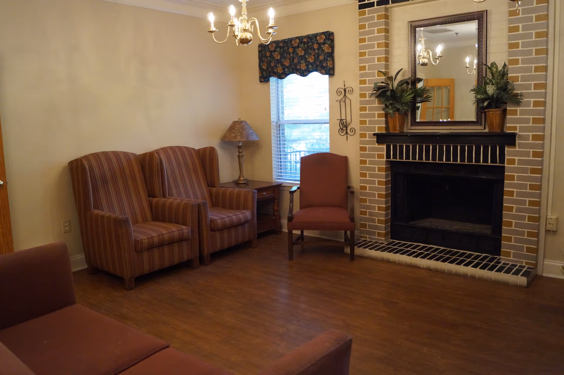 Living room with striped armchairs, a single upholstered chair by a window, and a brick fireplace topped with a mirror and potted plants.