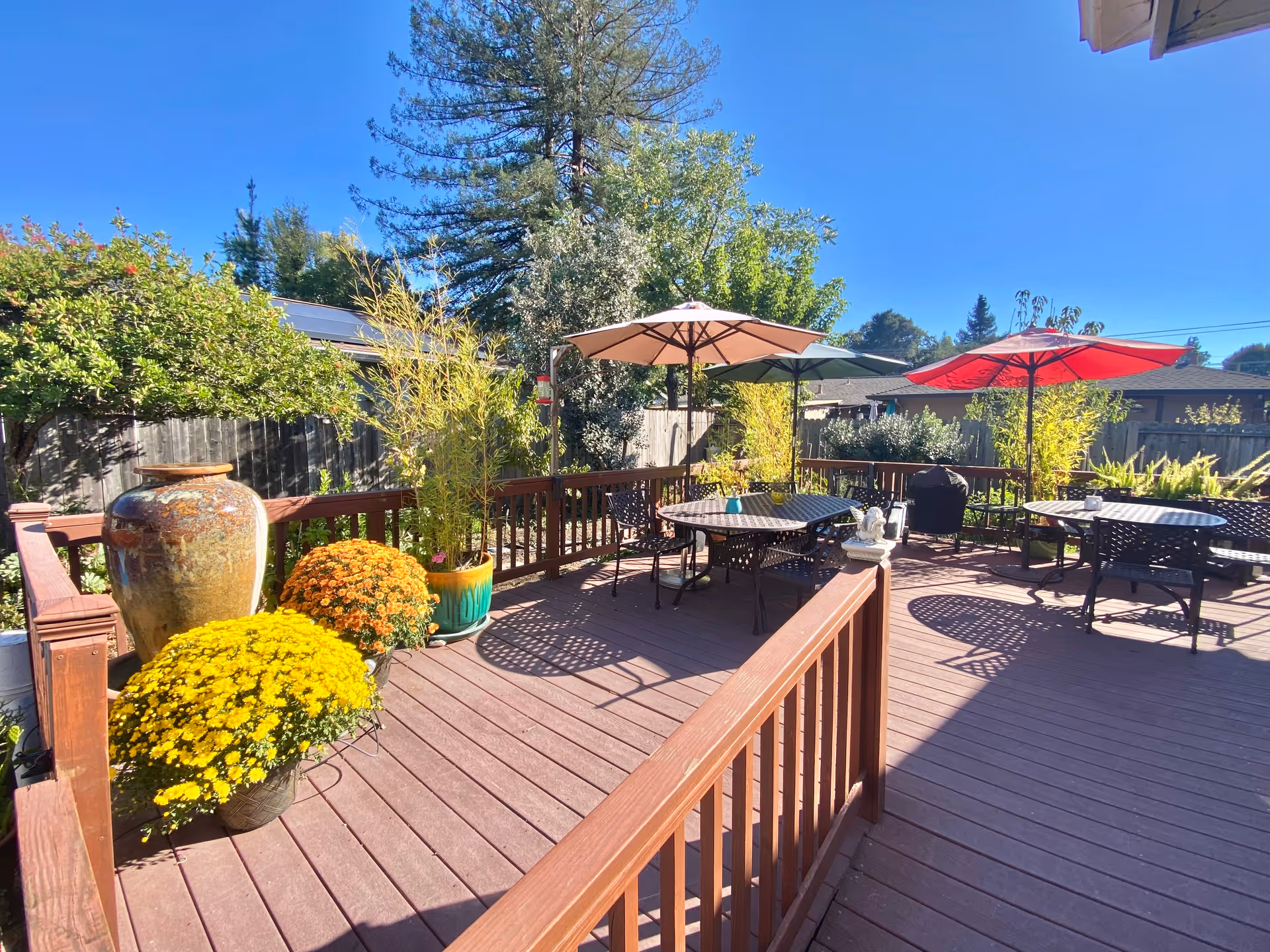 Outdoor wooden deck area with several round tables and chairs under large umbrellas. There are potted plants and flowers, including yellow and orange chrysanthemums, along the railing. Trees and a wooden fence surround the deck under a clear blue sky.