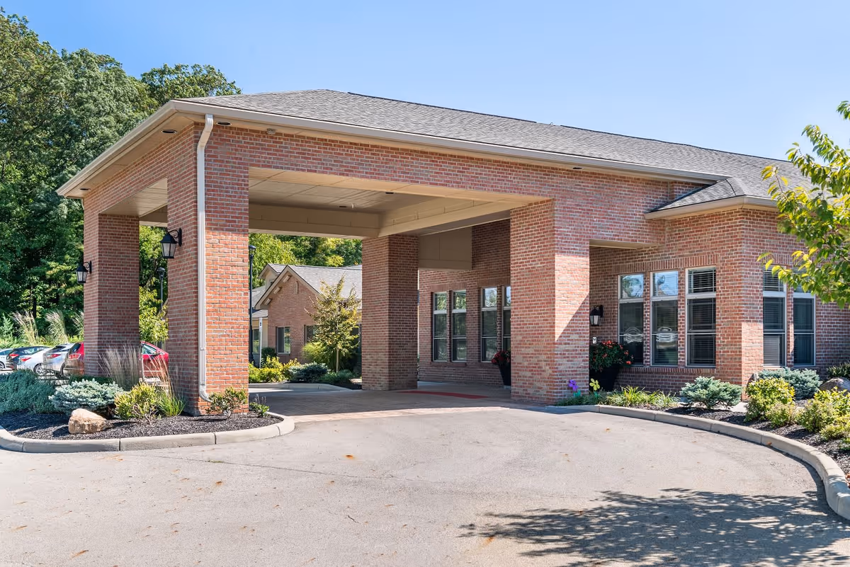 Front exterior view of a brick building with a covered driveway entrance, surrounded by landscaped greenery and parked cars under a clear blue sky.