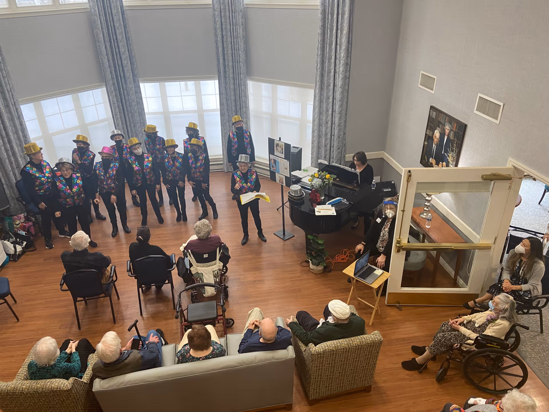 A group of elderly people seated in a common room watching a choir performance. The choir members are wearing colorful vests and shiny hats, some in gold and silver. A woman is playing the piano near the choir. The room has large windows with curtains and wooden flooring.