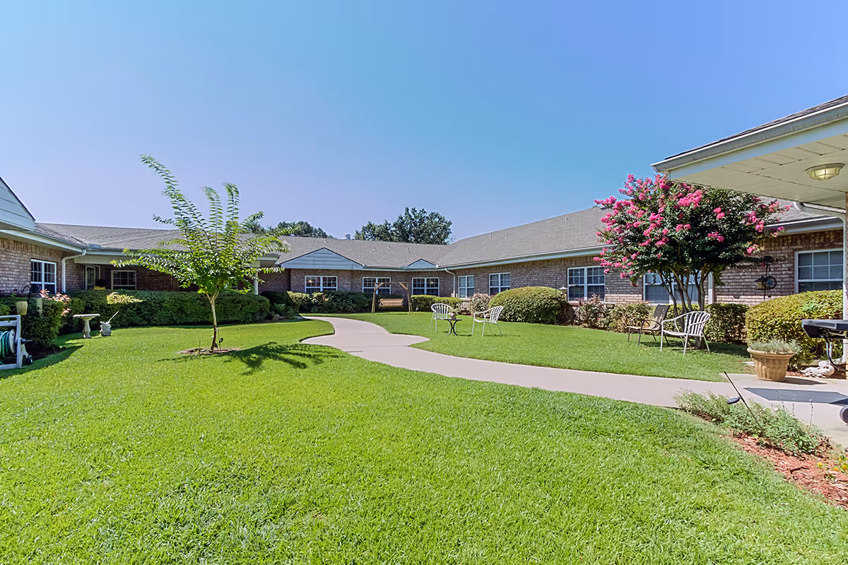 A well-maintained outdoor courtyard area at Alpine House with a green lawn, a paved walkway, several chairs and tables, a wooden swing, and flowering trees and shrubs surrounding a single-story brick building under a clear blue sky.