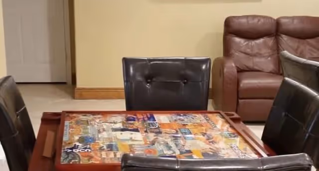 A small game table with a colorful puzzle or board on it surrounded by leather chairs and a brown recliner against a beige wall.