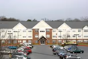 Front exterior view of a large three-story senior living facility named Sunnybrook Senior with a parking lot filled with cars in front. The building has a combination of brick and light-colored siding with multiple windows and a central entrance.