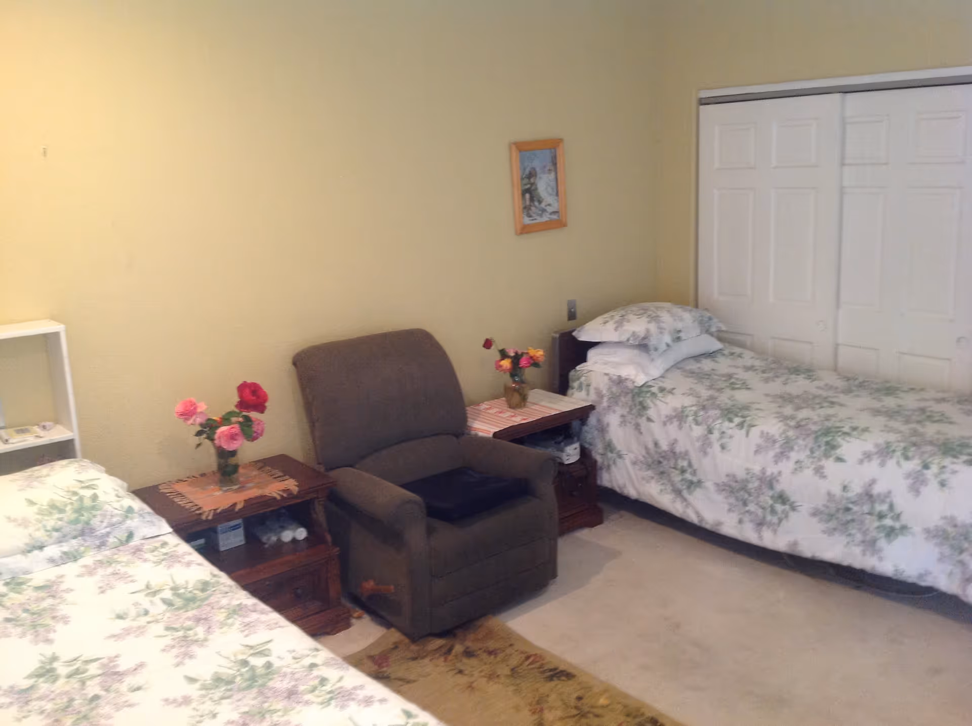A bedroom with two single beds covered in floral bedspreads, a brown recliner chair between the beds, two wooden nightstands with flower vases on top, a small white shelf, a framed picture on the wall, and a closed white closet door.