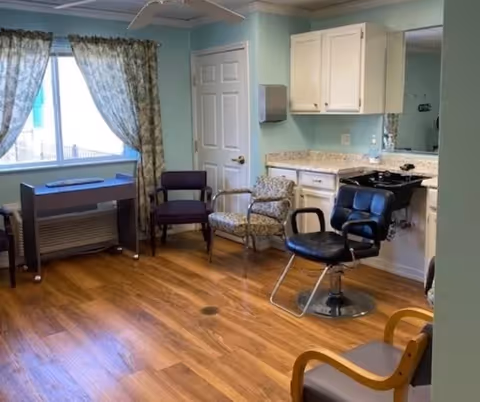 Interior room in an assisted living facility with salon styling chairs, a shampoo sink, cabinets, and several guest chairs by a curtained window.