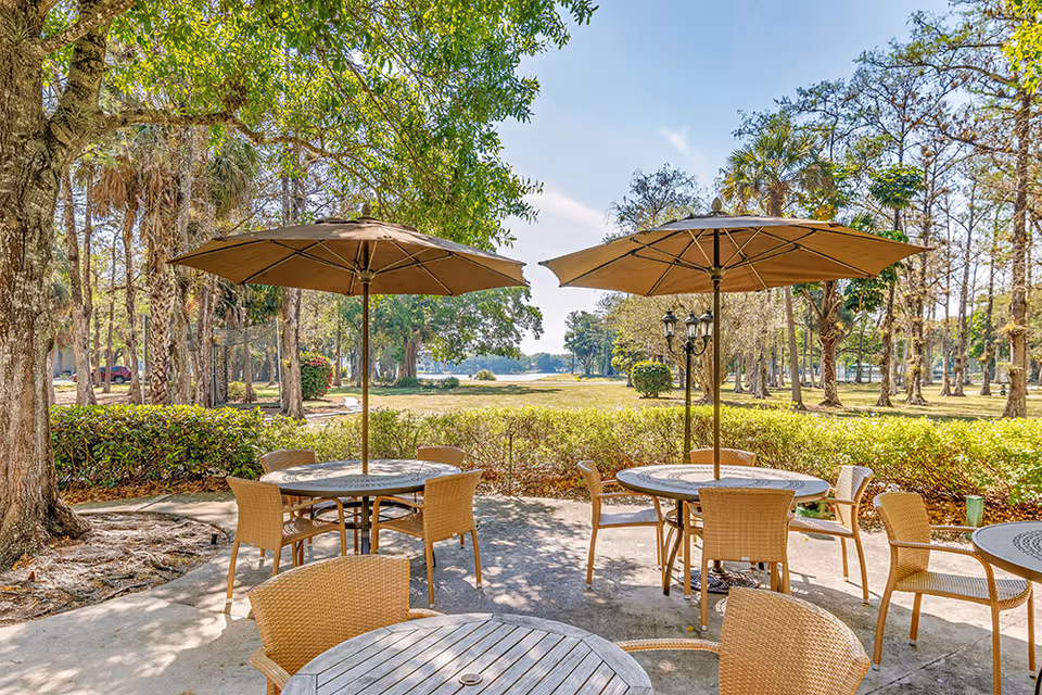 Patio with round tables, wicker chairs and large umbrellas overlooking a grassy, tree-lined outdoor area.