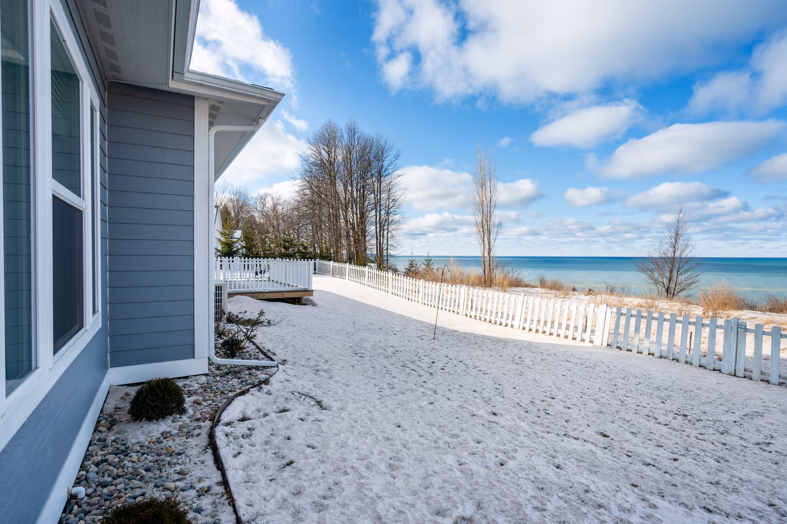 Snow-covered yard with a white picket fence next to a blue-gray building, leafless trees, and a view of a large body of water under a partly cloudy sky.