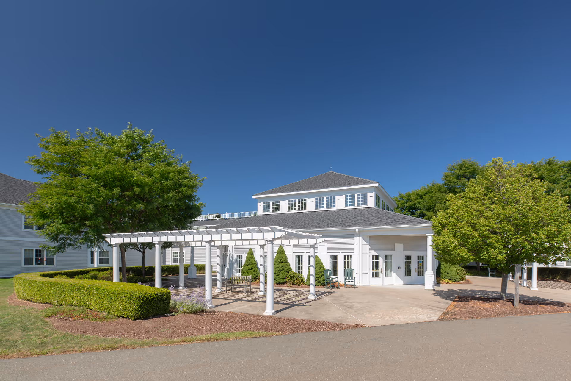 Exterior view of a white two-story building with a gray roof under a clear blue sky. The building has multiple windows and a covered patio area with white columns and benches. There are green trees and neatly trimmed bushes surrounding the building.