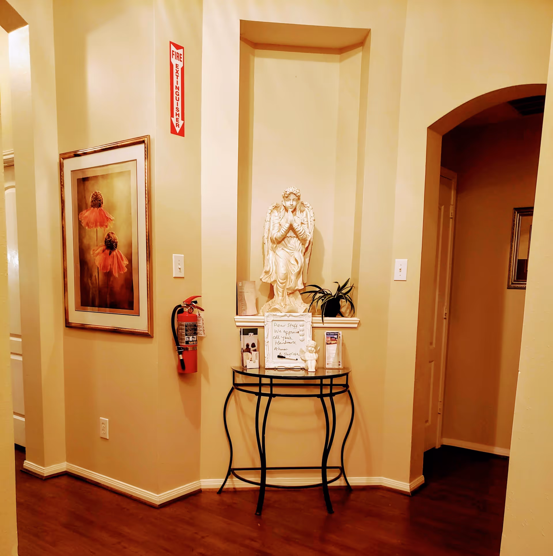 Interior corner of a facility with beige walls and wooden floor. A small black metal table with a glass top holds a white praying angel statue, a small potted plant, brochures, and a framed note. To the left, a fire extinguisher is mounted on the wall beneath a red fire extinguisher sign. A framed floral artwork hangs on the left wall. There is an arched doorway on the right side leading to another room.