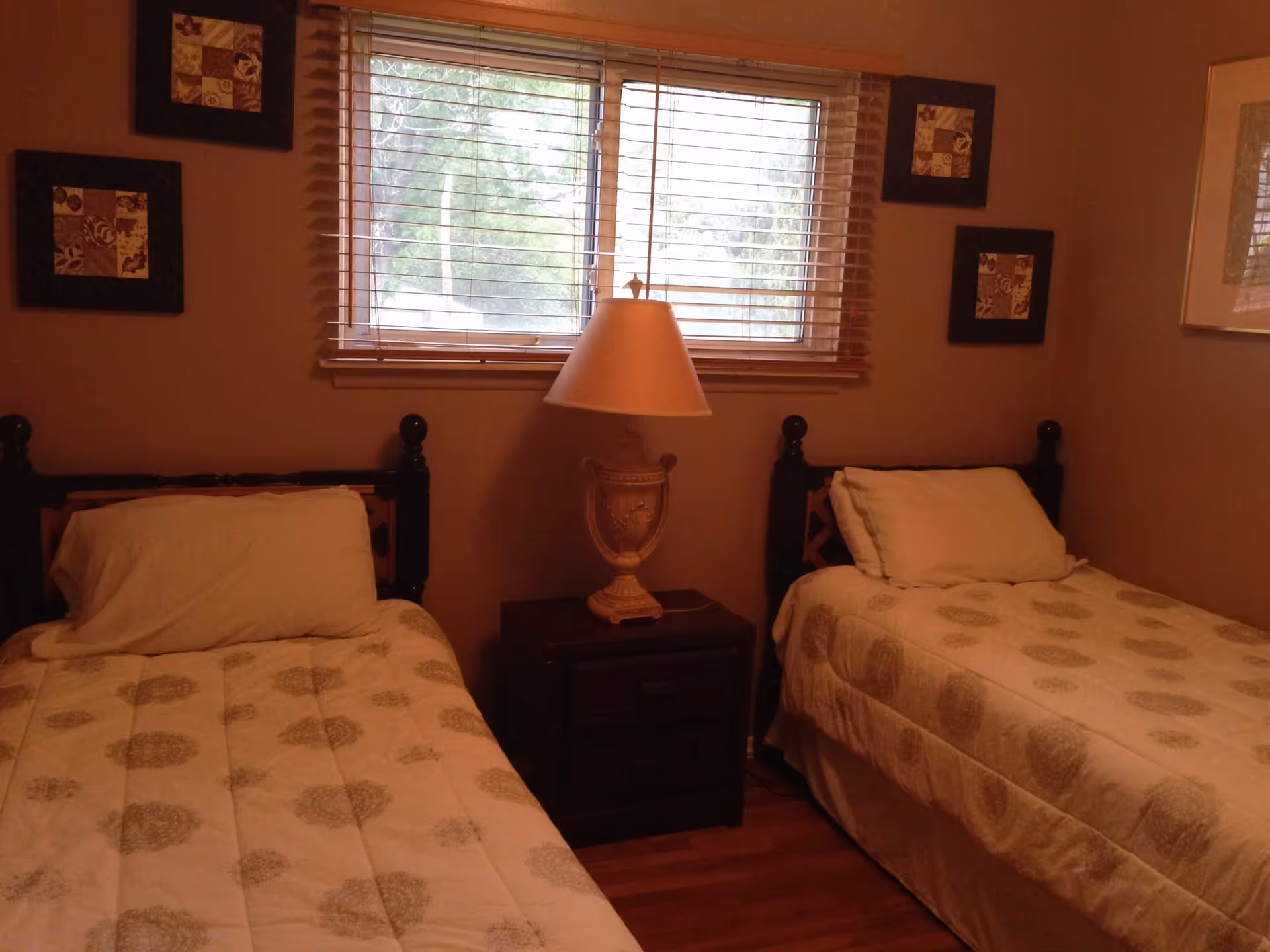 A bedroom with two twin beds separated by a dark wooden nightstand with a decorative lamp. The beds have matching beige comforters with a circular pattern and white pillows. A window with wooden blinds is centered above the nightstand, and framed artwork is hung on the walls around the window.