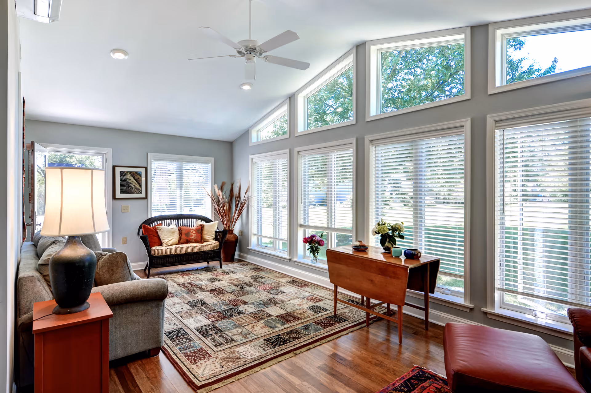 A bright living room with large windows featuring white blinds and triangular transom windows above. The room has a ceiling fan, a patterned area rug, a gray sofa with a side table and lamp, a wicker loveseat with cushions, a wooden drop-leaf table with decorative items, and a red leather ottoman. Outside greenery is visible through the windows.