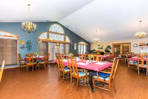A spacious dining room with multiple wooden tables and chairs arranged neatly. The tables are covered with red tablecloths and have small flower vases as centerpieces. The room features large windows with blinds and decorative stained glass above. There are chandeliers hanging from the ceiling, and the walls are painted blue and white. Various decorations and plants are placed around the room, creating a warm and inviting atmosphere.