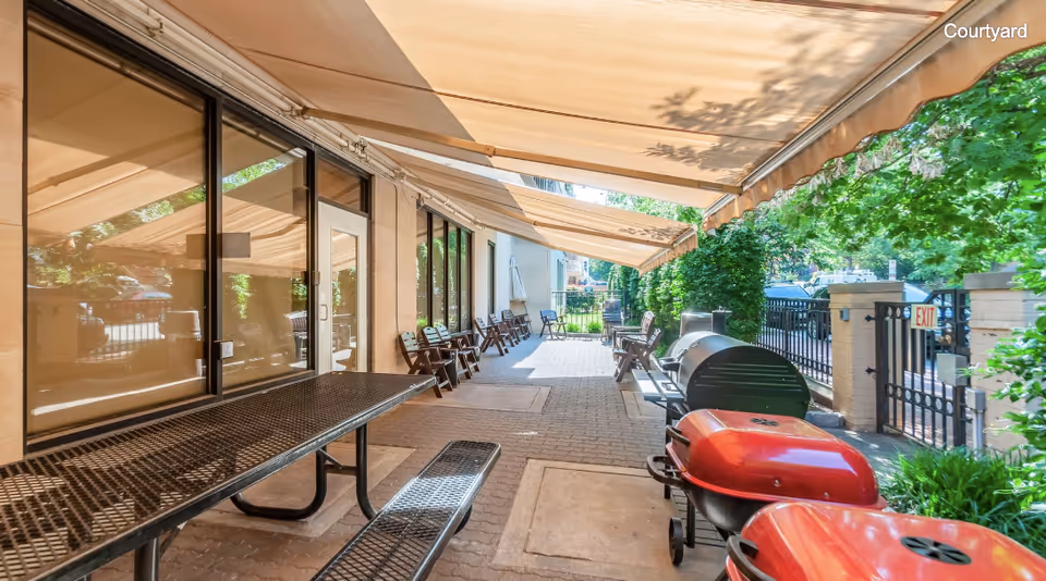 Outdoor courtyard area with multiple picnic tables and benches under large beige awnings. There are several chairs lined up against the building wall with large windows and a door. Two red grills and one black grill are visible on the right side near a black metal fence with greenery and trees providing shade.