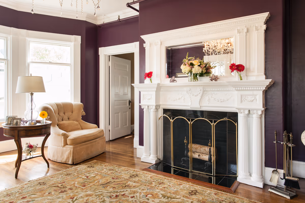A cozy living room with deep purple walls, a large ornate white fireplace with a gold fireplace screen, and a floral arrangement on the mantel. There is a beige upholstered armchair next to a wooden side table with a lamp and a small flower vase. A patterned area rug covers the wooden floor, and sunlight streams in through large windows.