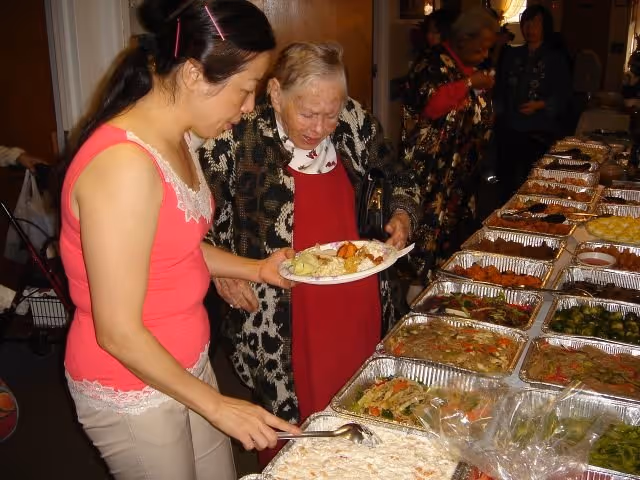 Two women serving themselves food from a buffet table filled with various dishes in aluminum trays. One woman is wearing a pink sleeveless top and the other is an elderly woman in a patterned cardigan and red dress. Other people are visible in the background.