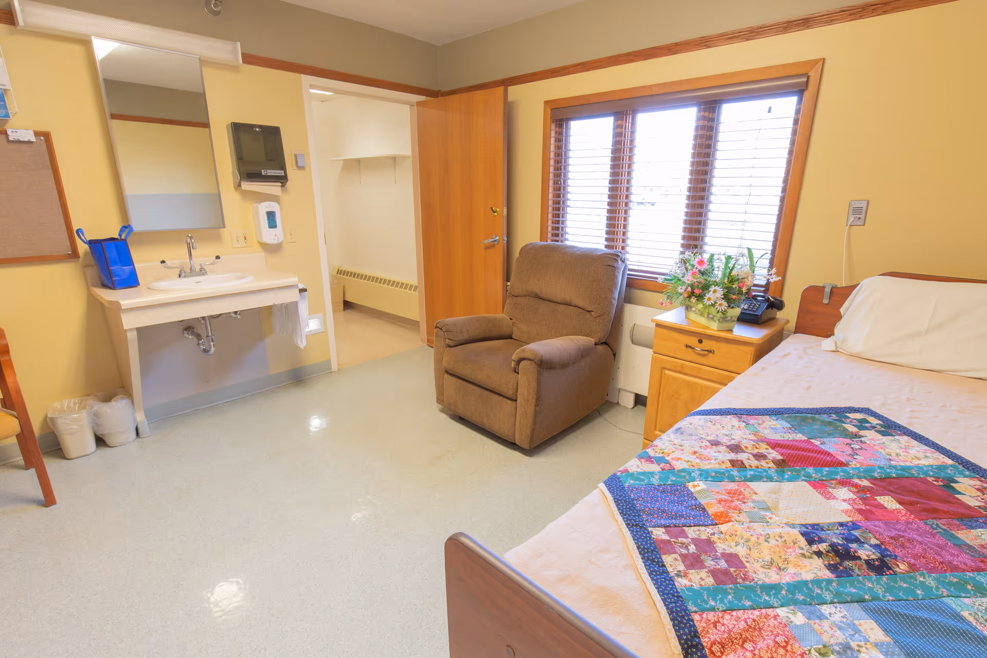 Resident bedroom with a bed covered by a colorful quilt, a recliner by a window, and a wall-mounted sink area.
