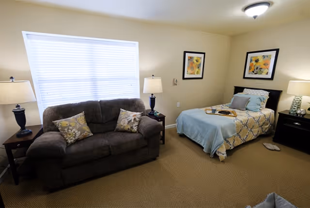 A cozy room with a dark brown sofa adorned with two floral pillows, a single bed with a blue and yellow patterned bedspread and multiple pillows, two bedside tables each with a lamp, two framed floral artworks on the wall, and a window with closed blinds letting in natural light.