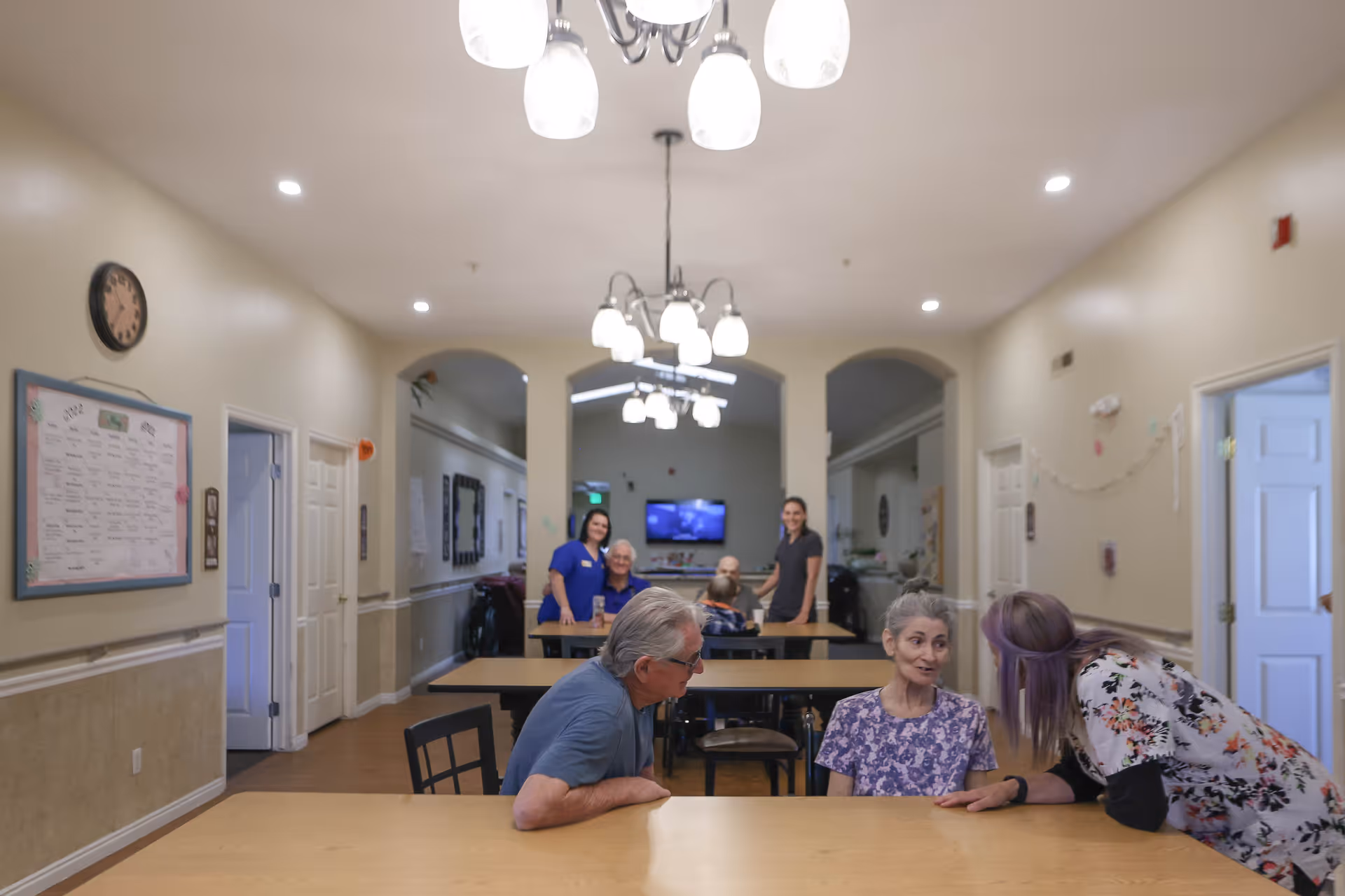 Residents and staff interact around tables in a brightly lit communal dining room of a memory care facility.