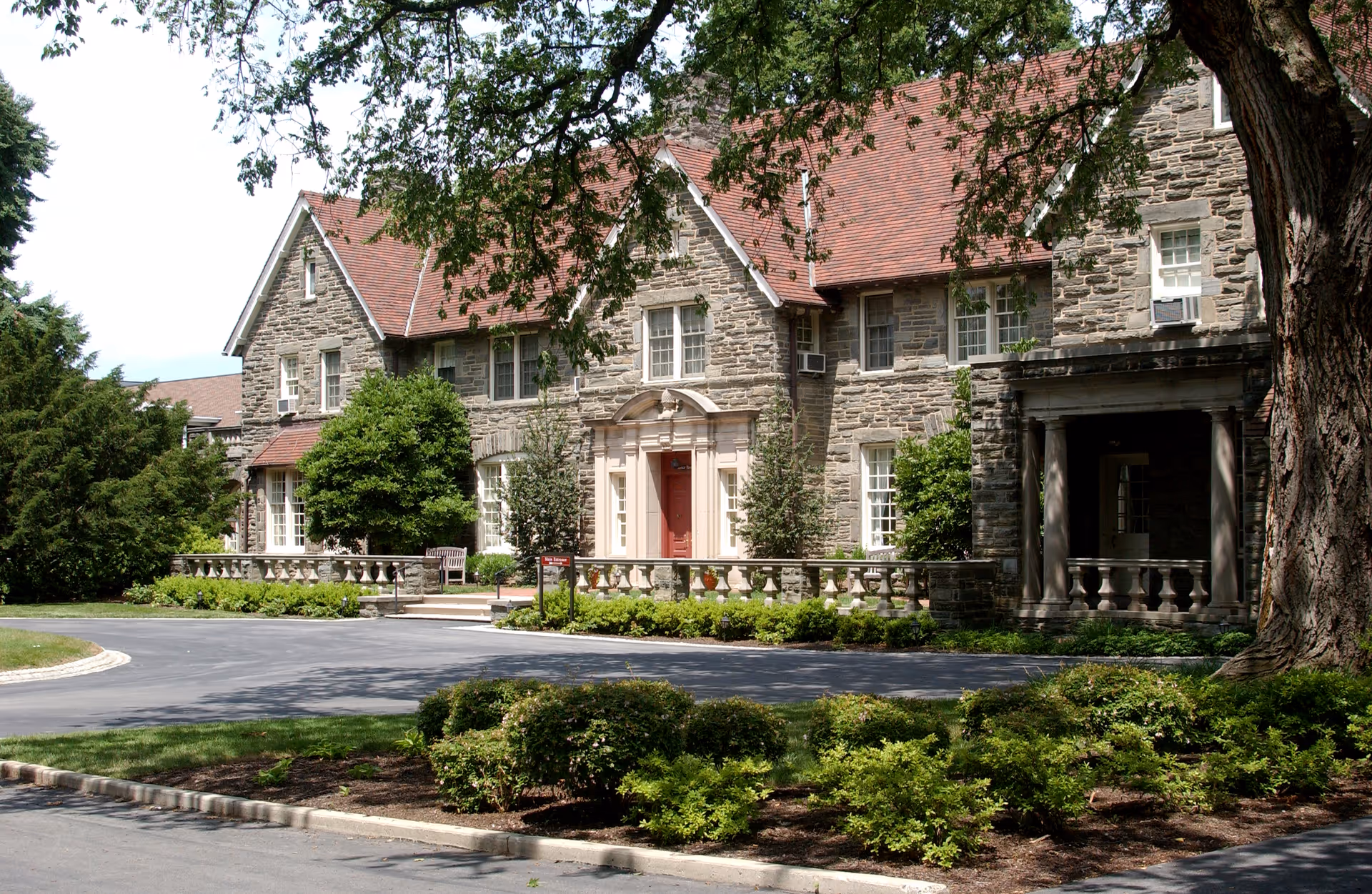 Front view of a stone manor-style building with a red roof, central entrance, and landscaped driveway.