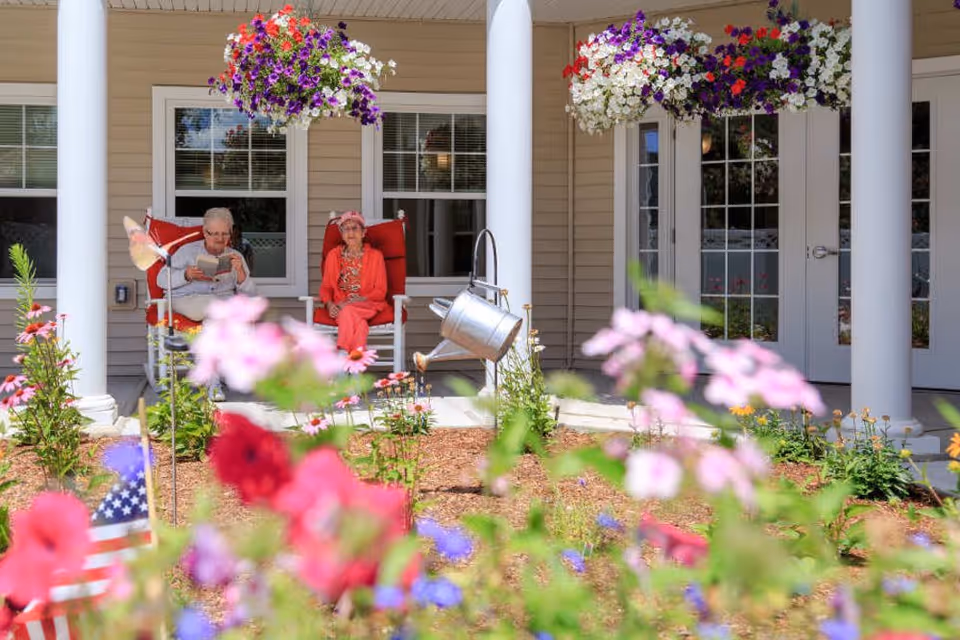 Two elderly women sitting on red cushioned rocking chairs on a porch of a building, with colorful hanging flower baskets above them and a garden with various flowers and an American flag in the foreground.