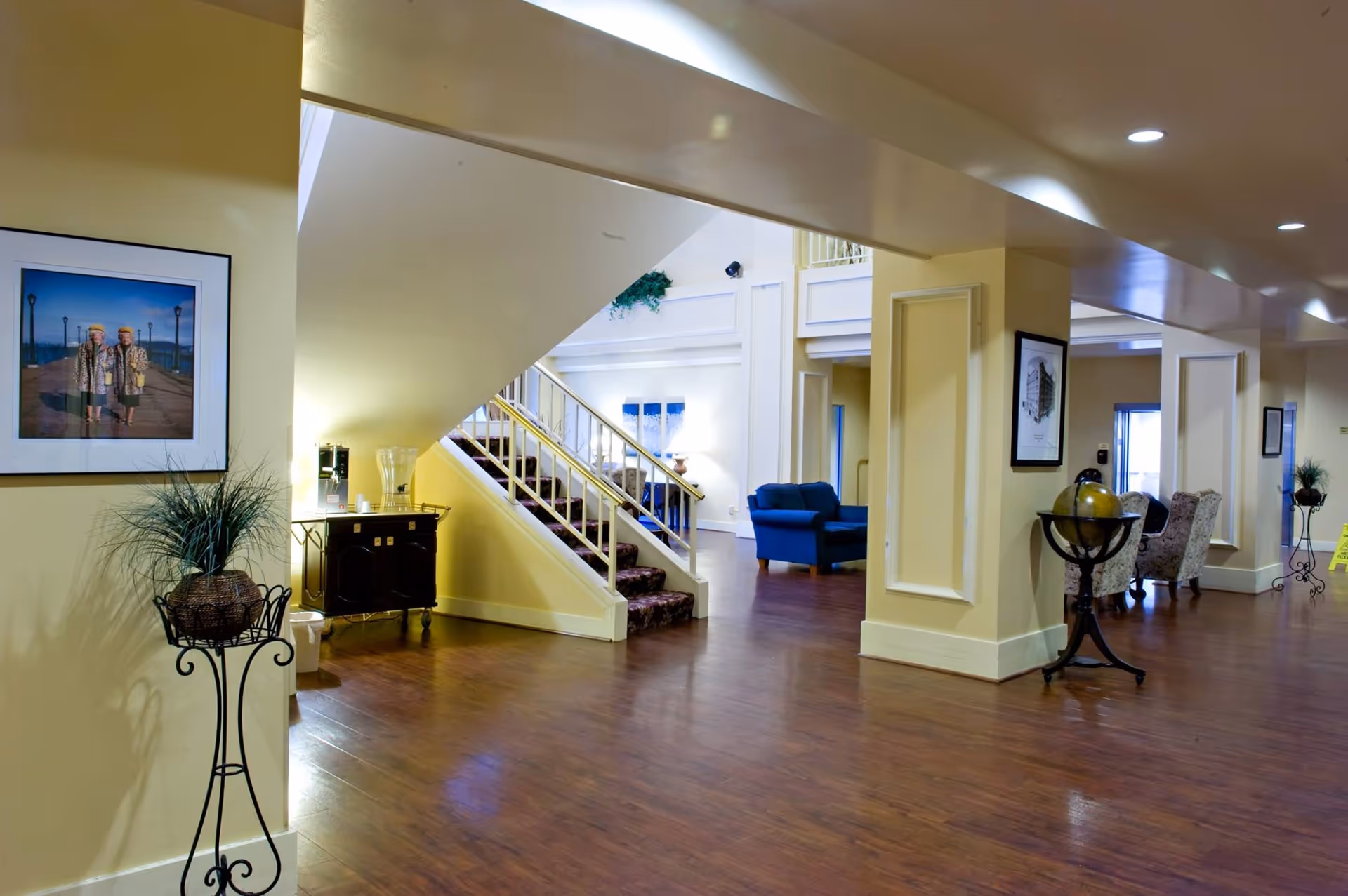 Interior view of a senior living community common area with wooden flooring, beige walls, a staircase with carpeted steps, framed pictures on the walls, a blue armchair, decorative plants, and a globe on a stand.