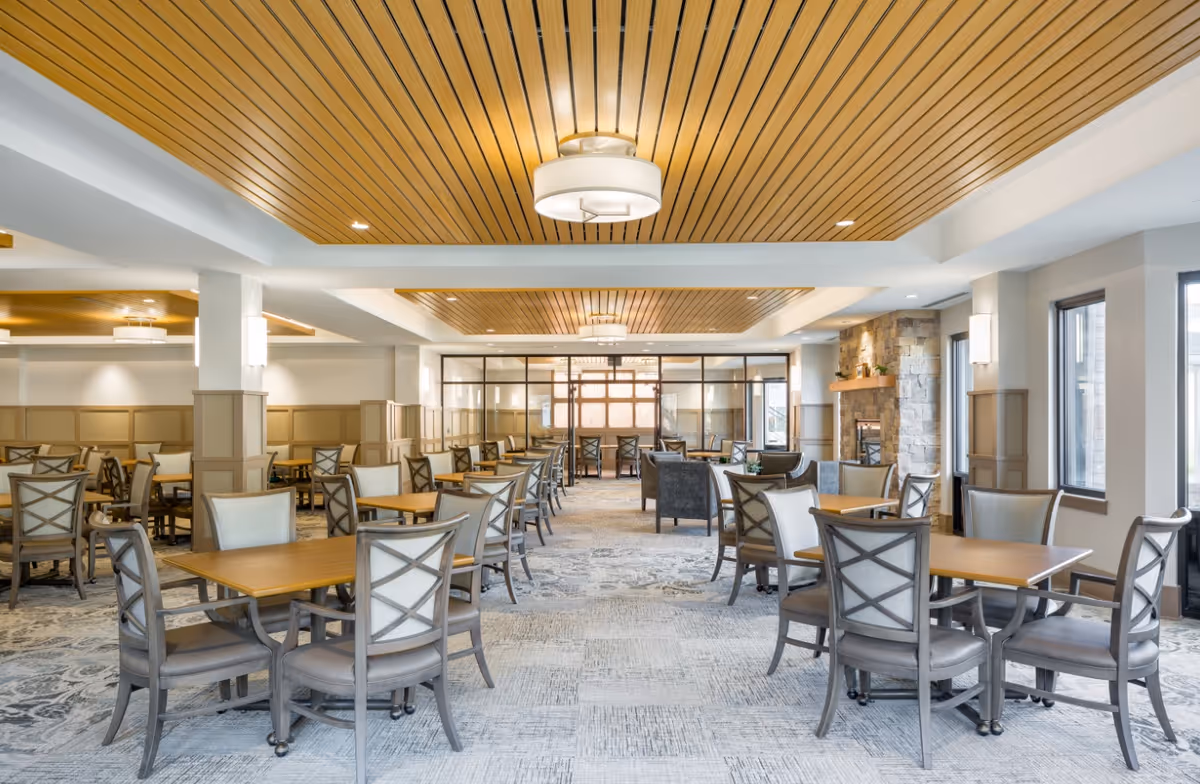 A spacious and well-lit dining room with multiple wooden tables and cushioned chairs arranged neatly. The room features a wooden slatted ceiling with modern light fixtures, large windows allowing natural light, and a stone fireplace on the right side. The flooring has a patterned carpet, and the walls are painted in neutral tones with wainscoting.