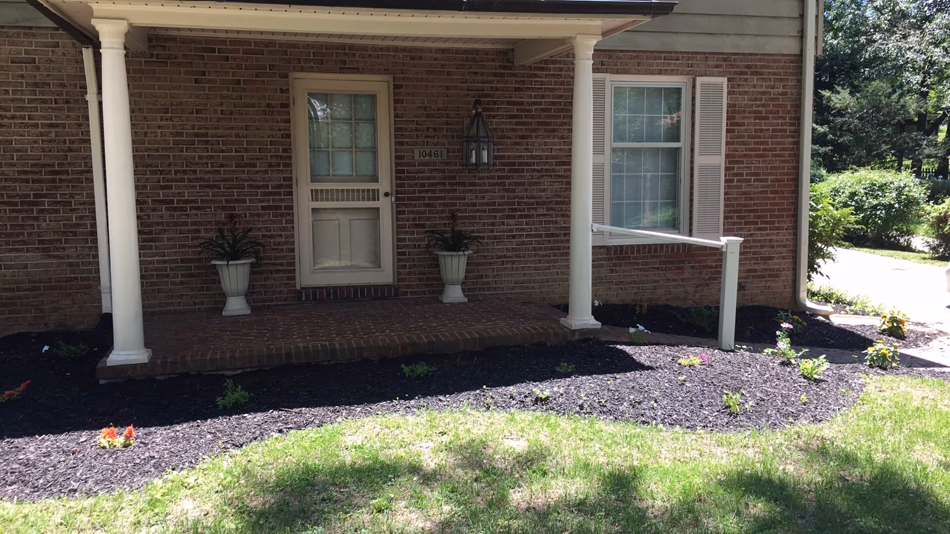 Front entrance of a brick building with a small porch supported by two white columns. There is a white door with a window and a house number plaque displaying 10461. Two potted plants are placed symmetrically on either side of the door. The porch is surrounded by a garden bed with dark mulch and small plants, and a grassy lawn is visible in the foreground.